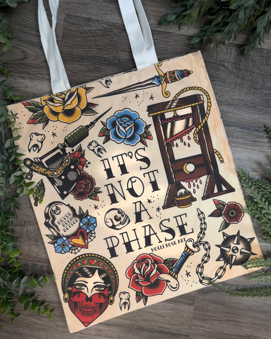Tote bag with tattoo-style design and text 'It's Not a Phase' on a wooden surface with plants.