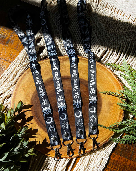 a black lanyard with the words be weird stay spooky and white illustrations