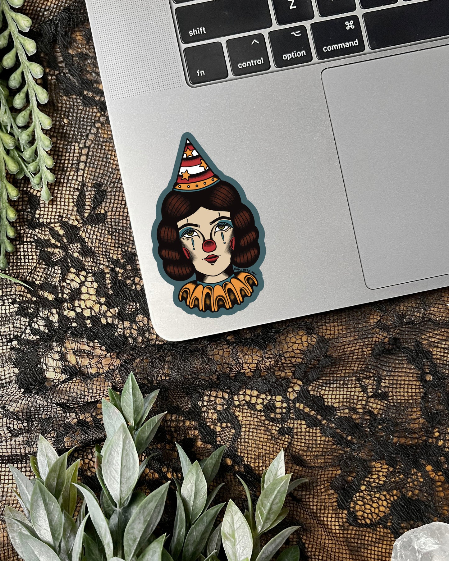 Laptop with a sticker of a clowns face on a lace surface with plants.