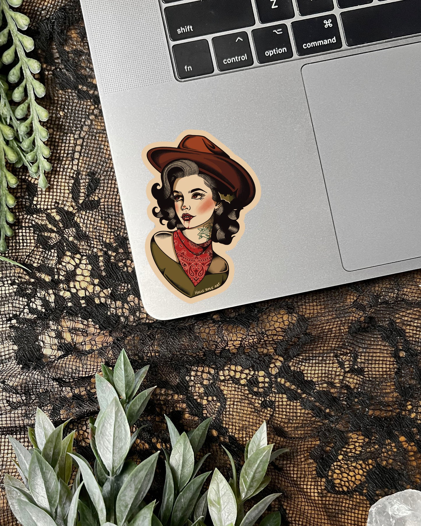 Laptop with a decorative sticker of a vampire cowgirl wearing a hat and scarf on a lace surface.