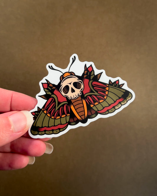 Sticker with a skull and moth design held by a hand against a brown background