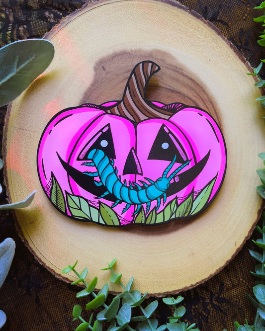 Hand holding a pink pumpkin with a cartoonish face and green worm design against a white background