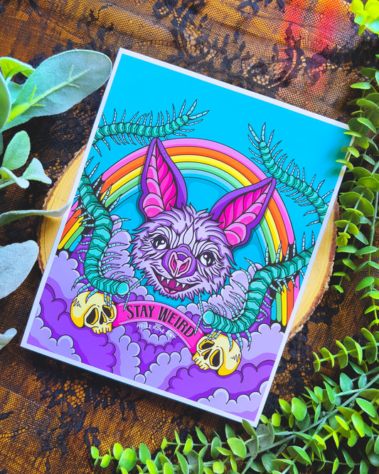 an art print with a purple bat face, a rainbow and the words stay weird at the bottom with skulls