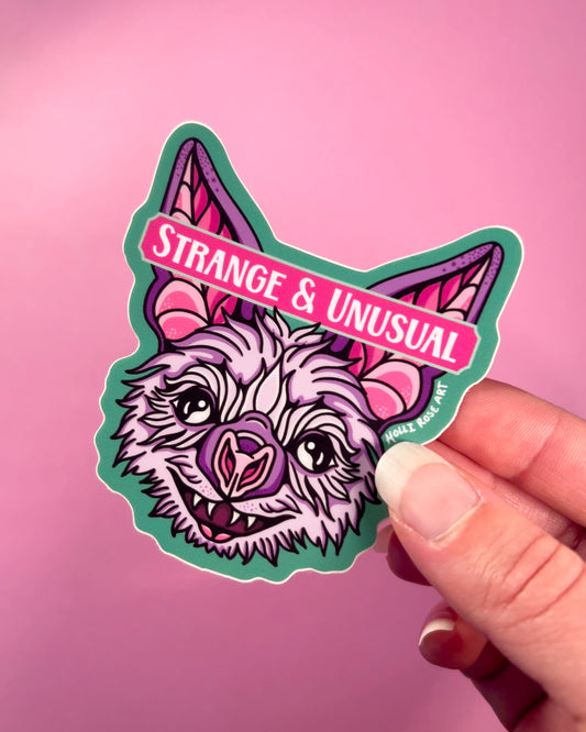 Sticker with a bat design and 'Strange & Unusual' text held by a hand against a pink background