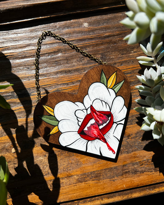 A painting on a small wooden heart of a split tongue coming out of a white flower