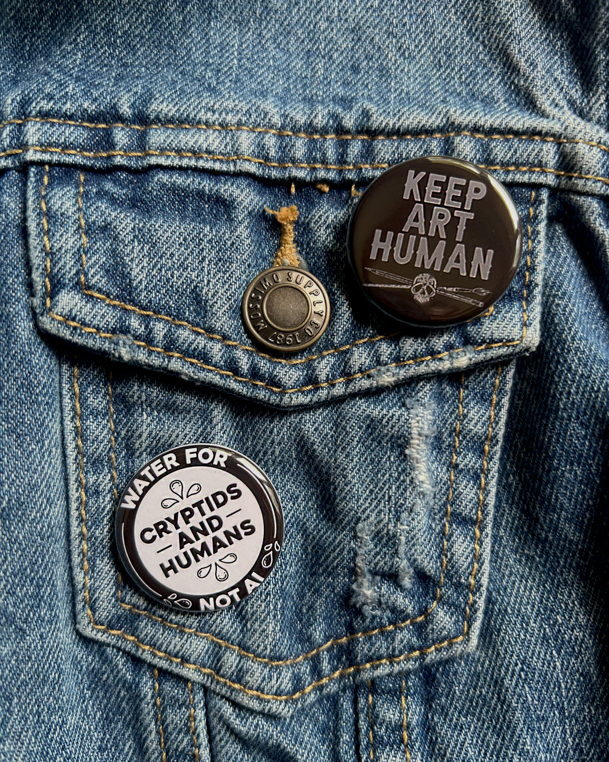 2 pins on a jean pocket - one that says keep art human and the other that says water for cyptids and humans not ai