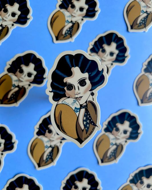 A sticker with a pinup style gal with blue hair, button eyes and a yellow shirt
