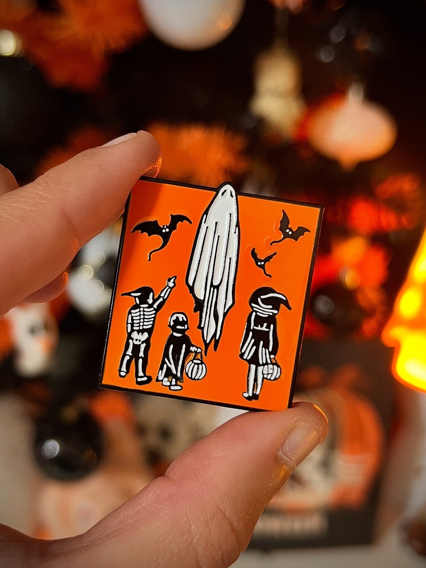 Halloween-themed badge with ghost, witch, and bat design held by a hand against a blurred festive background.