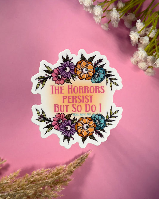 Sticker with floral design and text  saying "the horrors persist but so do I" on a pink background with flowers
