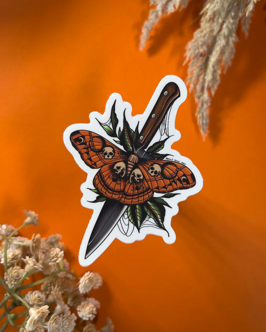 A sticker with a moth that as skulls on it and a knife behind it 