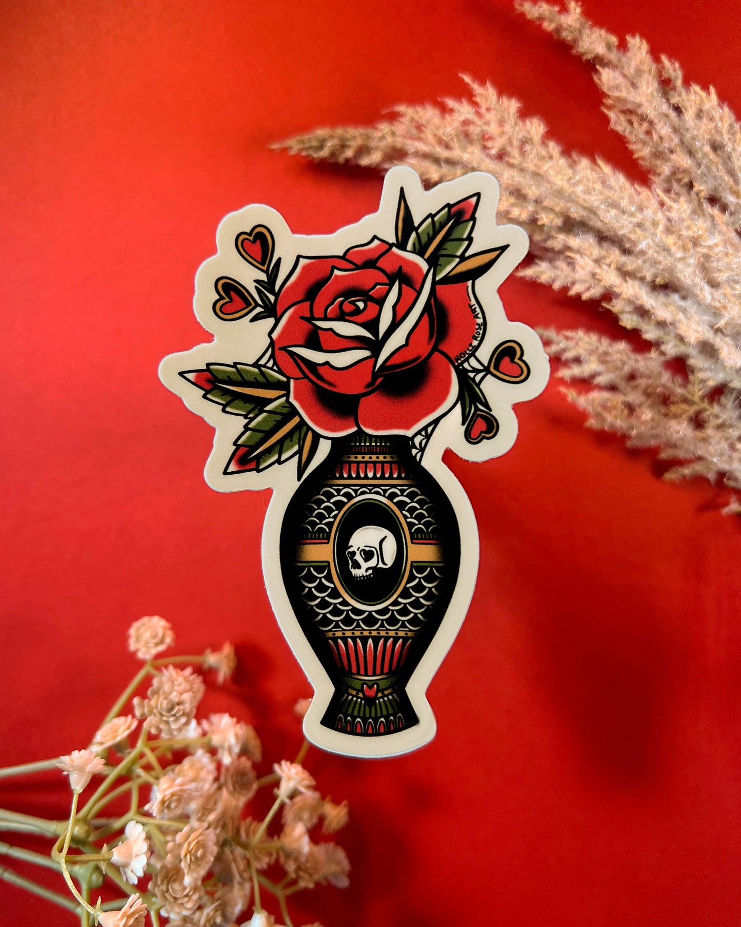 Decorative sticker with a rose and skull design on a red background with dried flowers.