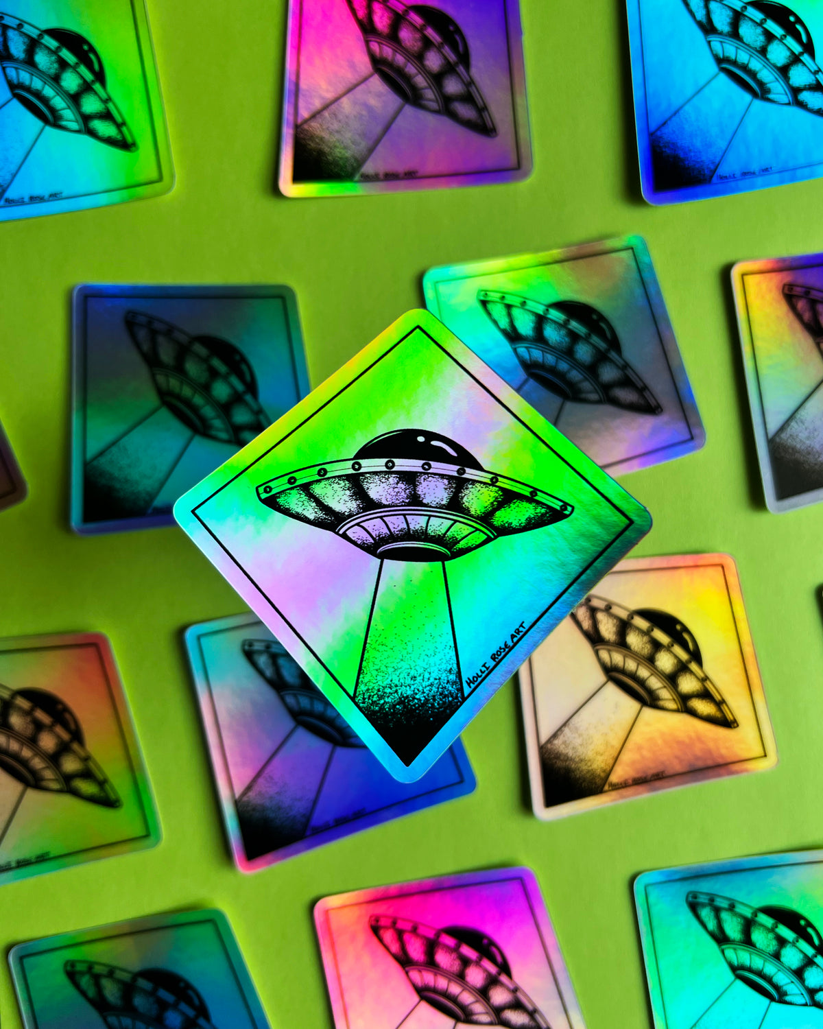 A diamond-shaped holographic sticker featuring a black line art drawing of a UFO, displayed on a bright lime green background with other matching stickers scattered around.
