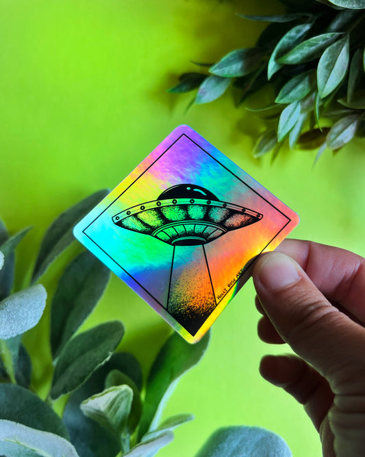 A hand holds a diamond-shaped holographic sticker featuring a black UFO graphic, set against a bright lime green background with green leaves.