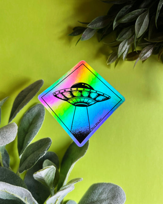 A diamond-shaped holographic sticker featuring a black UFO graphic, displayed on a bright lime green background with decorative green leaves.