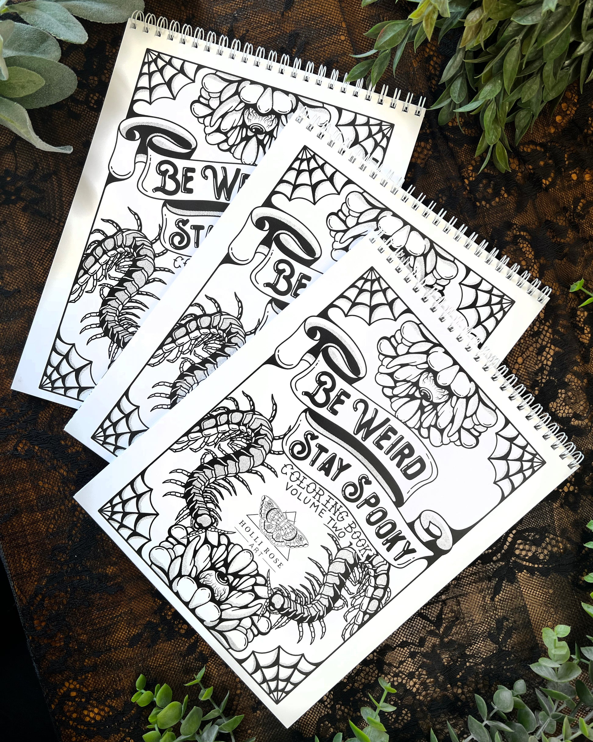 A spiral-bound coloring book with black and white Halloween-themed illustrations, titled 'Be Weird Stay Spooky Coloring Book Vol. 2'.