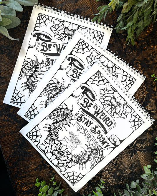 A spiral-bound coloring book with black and white Halloween-themed illustrations, titled 'Be Weird Stay Spooky Coloring Book Vol. 2'.