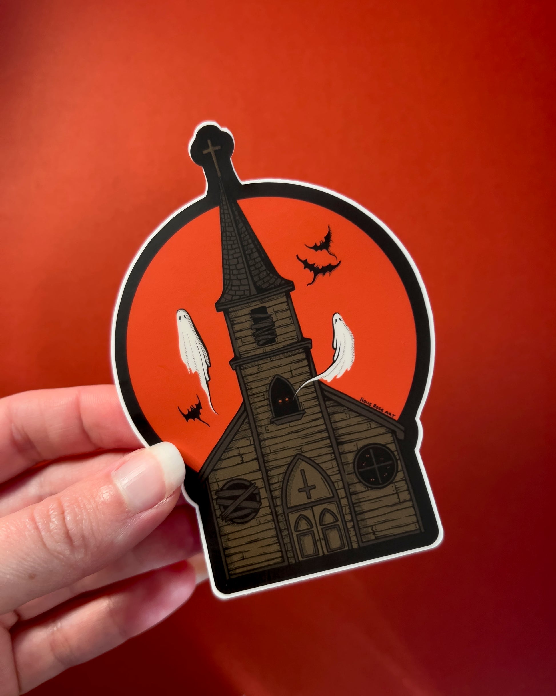 A sticker of a haunted church with ghosts and bats flying around and a red moon behind it