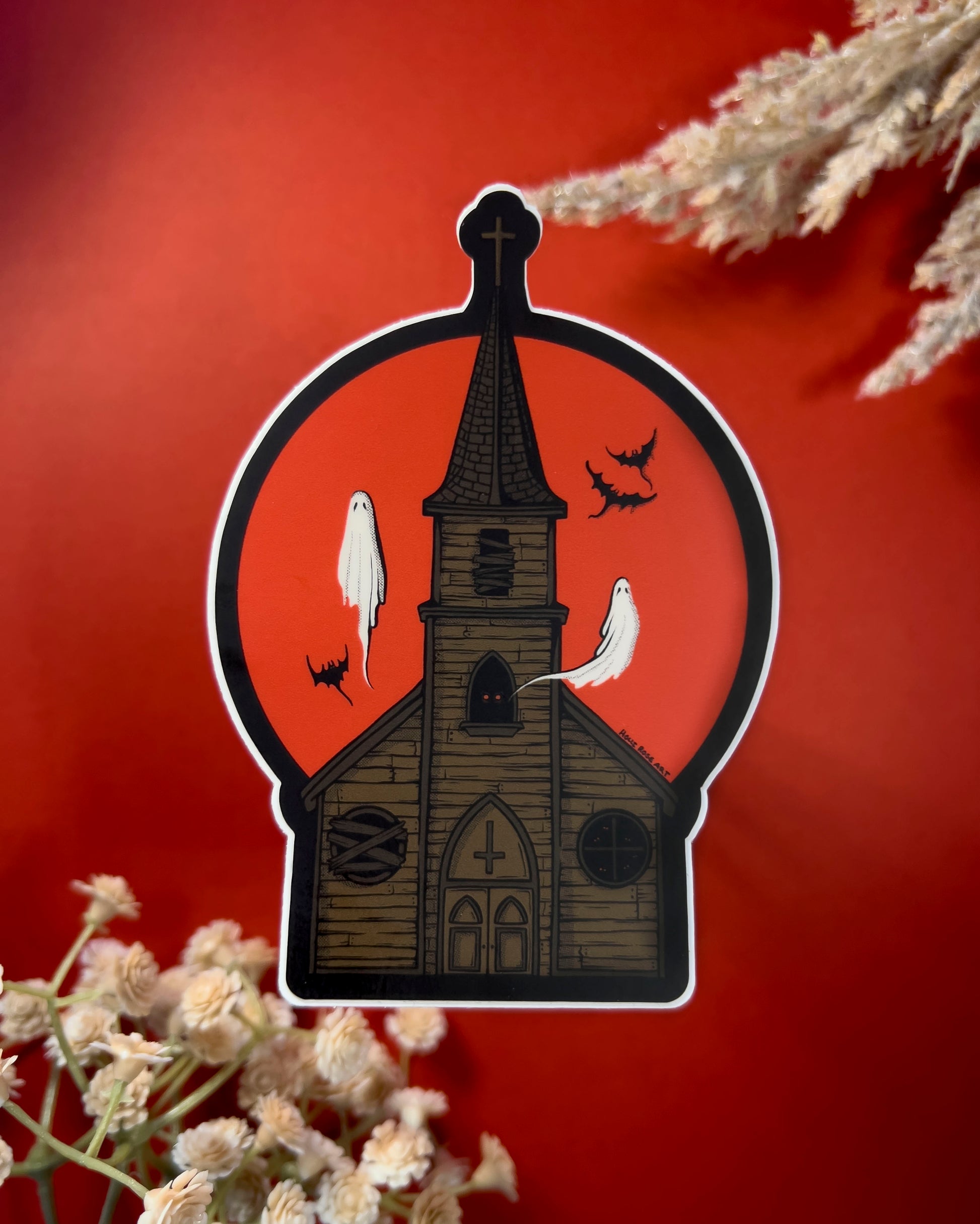 A sticker of a haunted church with ghosts and bats flying around and a red moon behind it