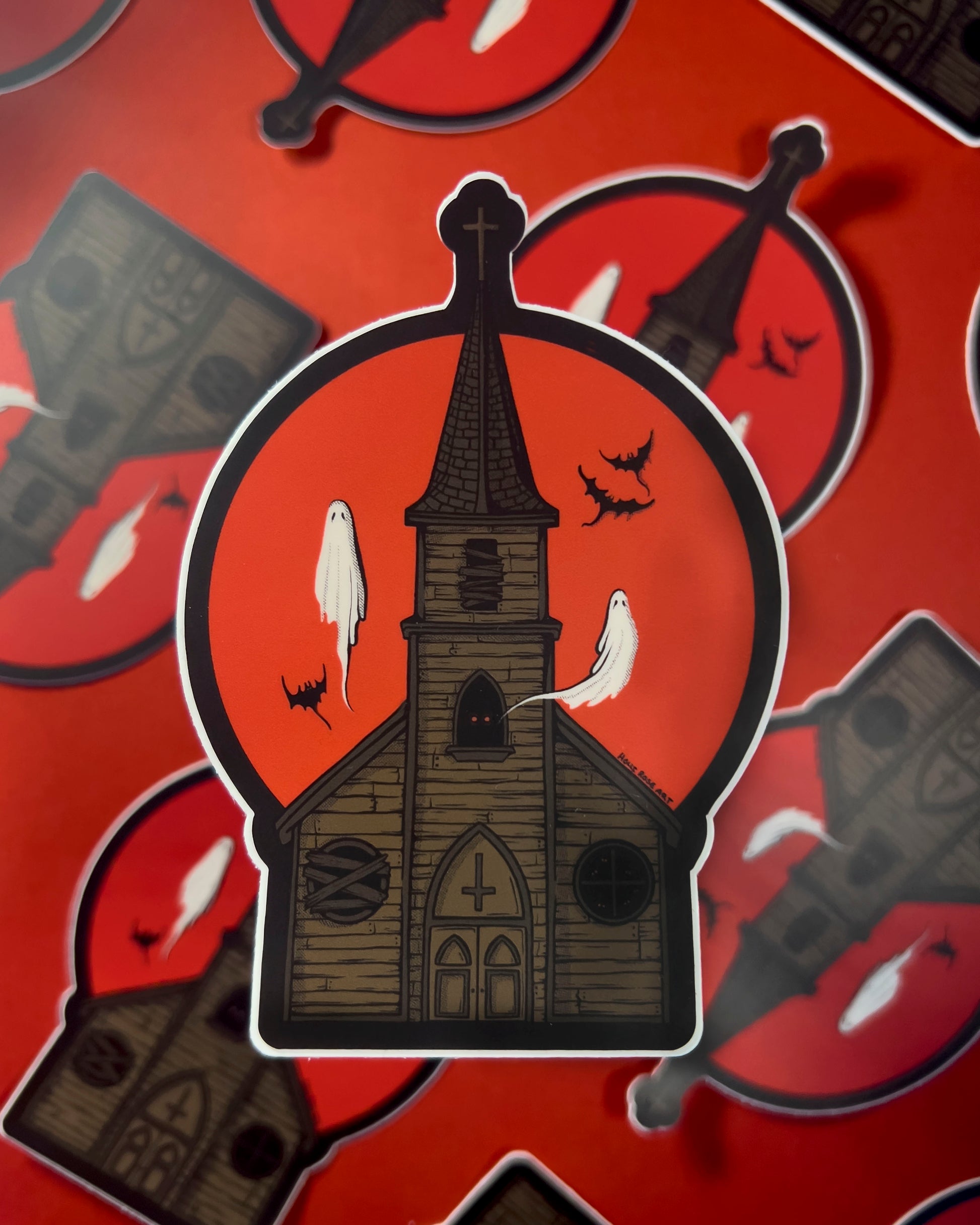 A sticker of a haunted church with ghosts and bats flying around and a red moon behind it