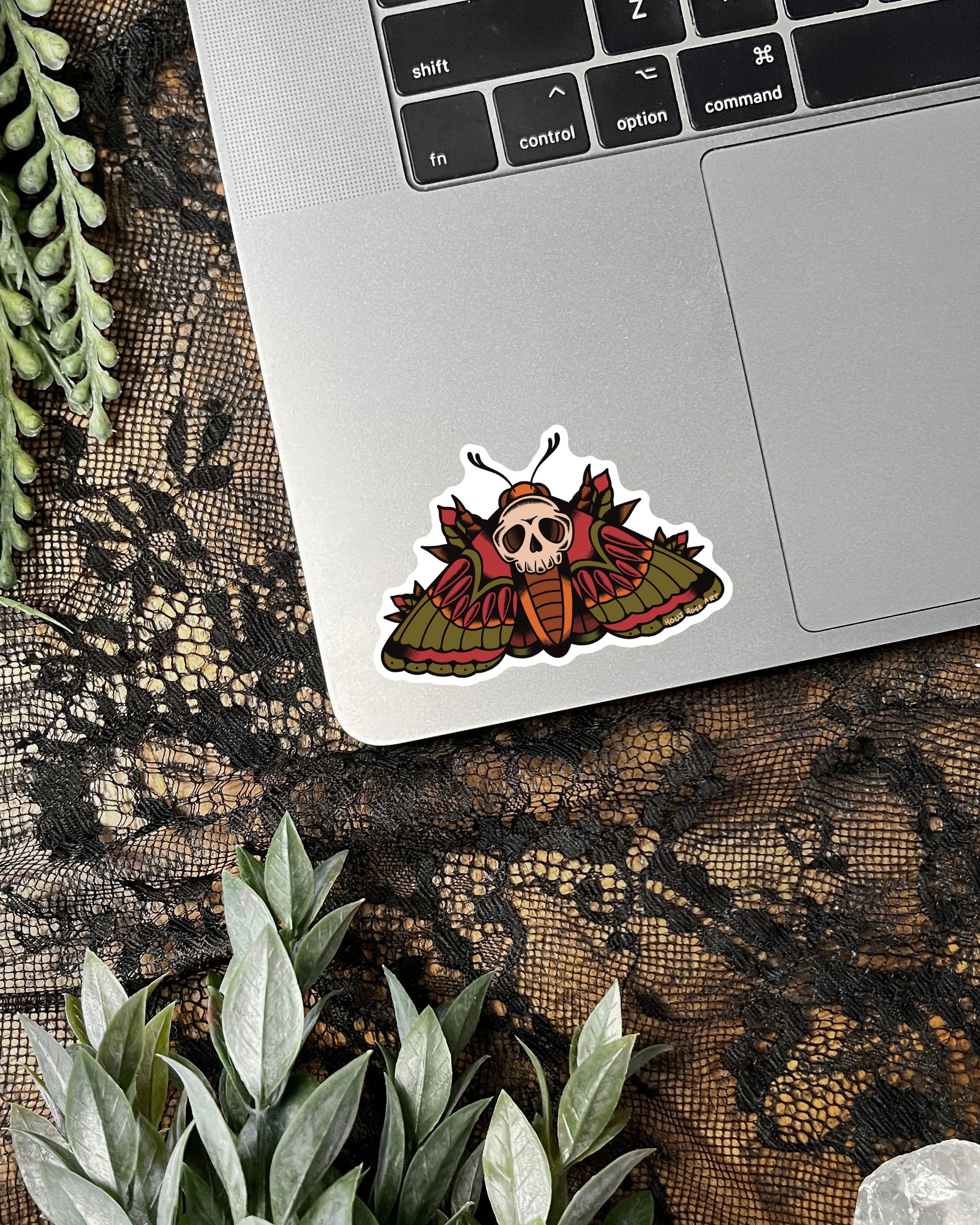 Sticker with a skull moth design on a laptop keyboard surrounded by lace and plants