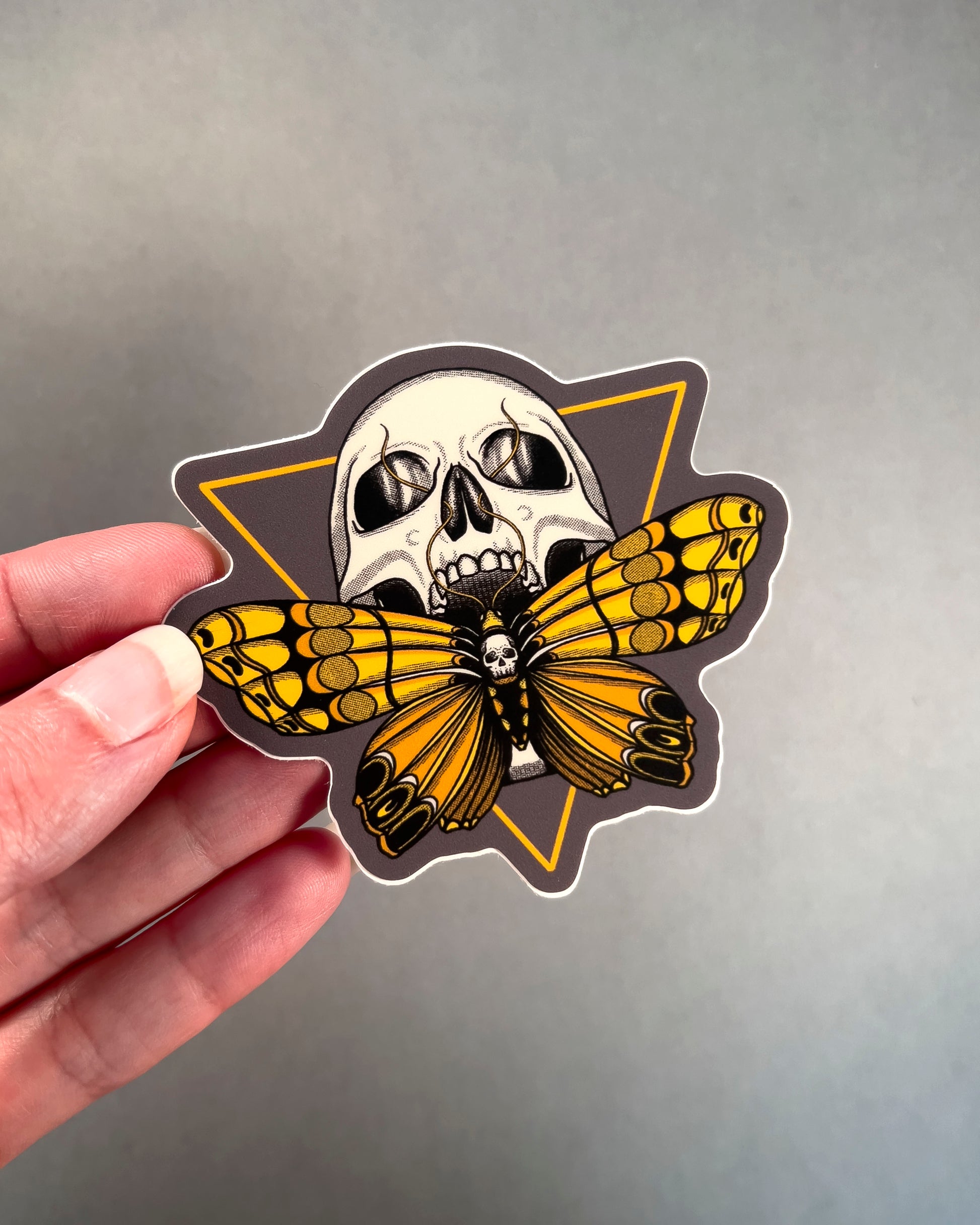 Sticker with a skull and butterfly design held by a hand on a gray background