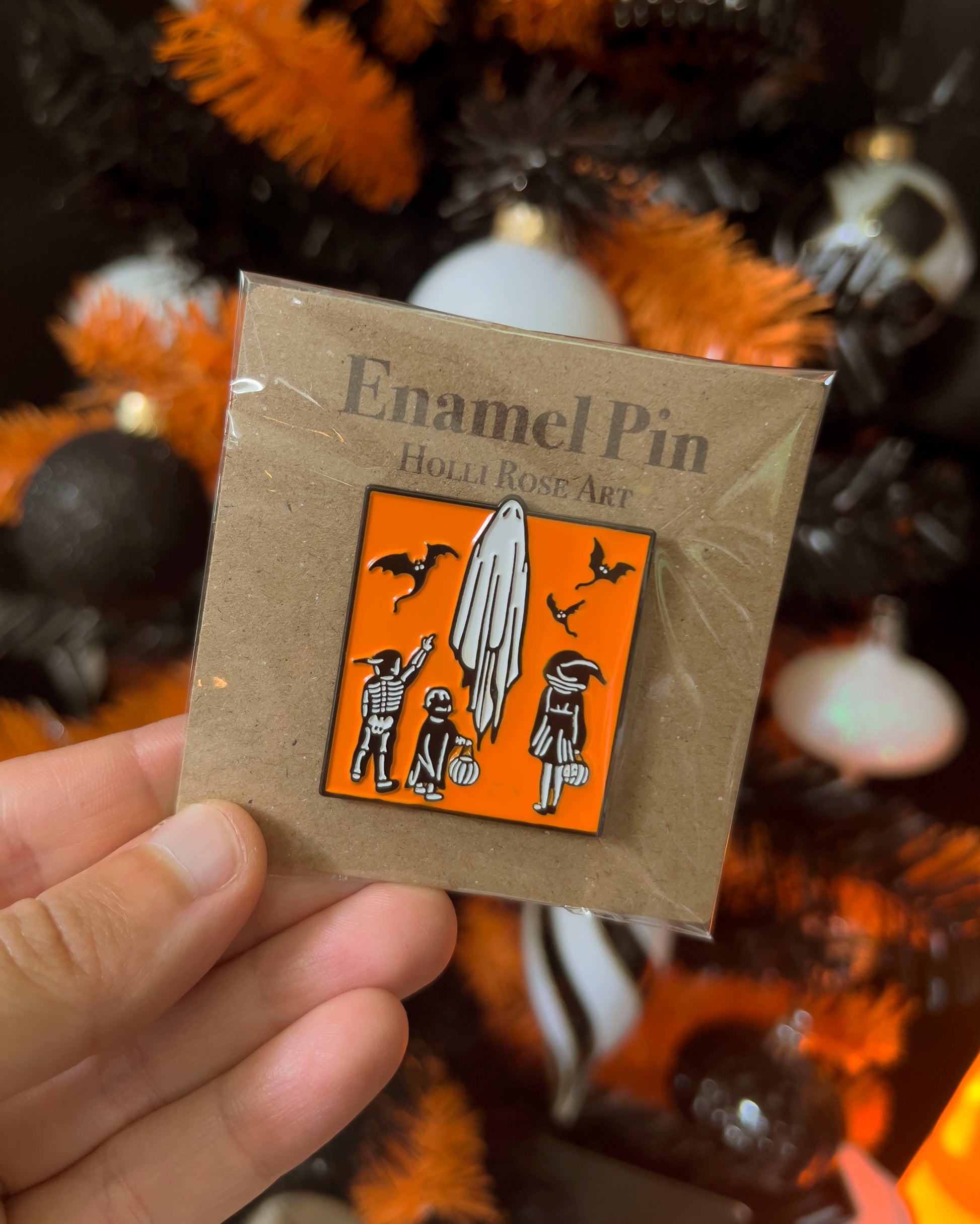 Enamel pin with ghost design on a brown card, held in front of Halloween-themed decorations.
