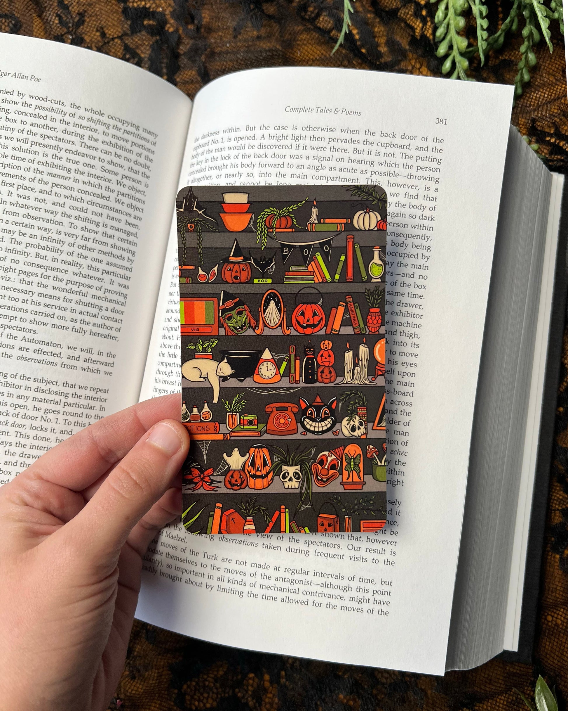 A hand holding a Halloween-themed bookmark with various icons and text, placed within an open book.