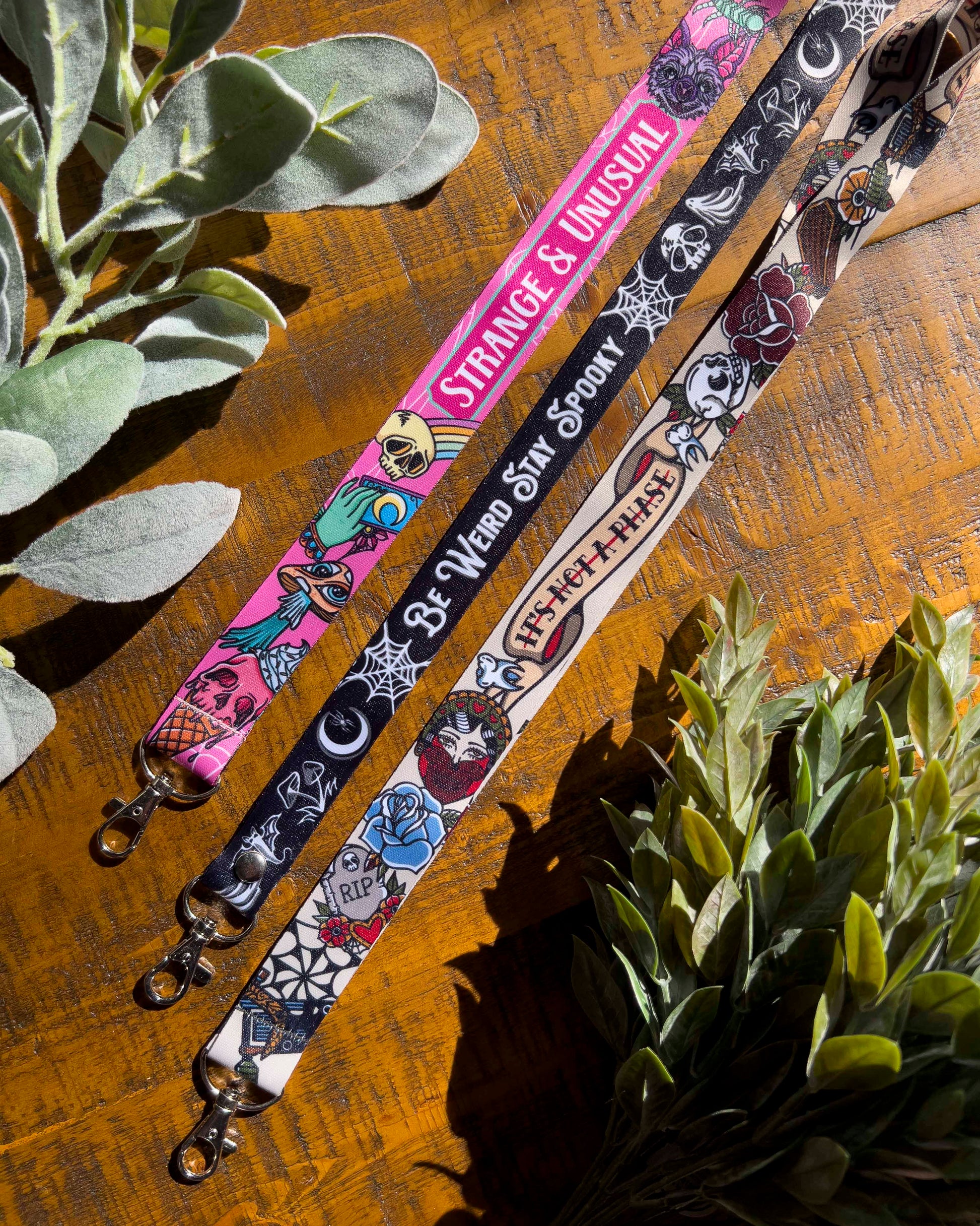 Three colorful lanyards with unique designs on a wooden surface with plants.