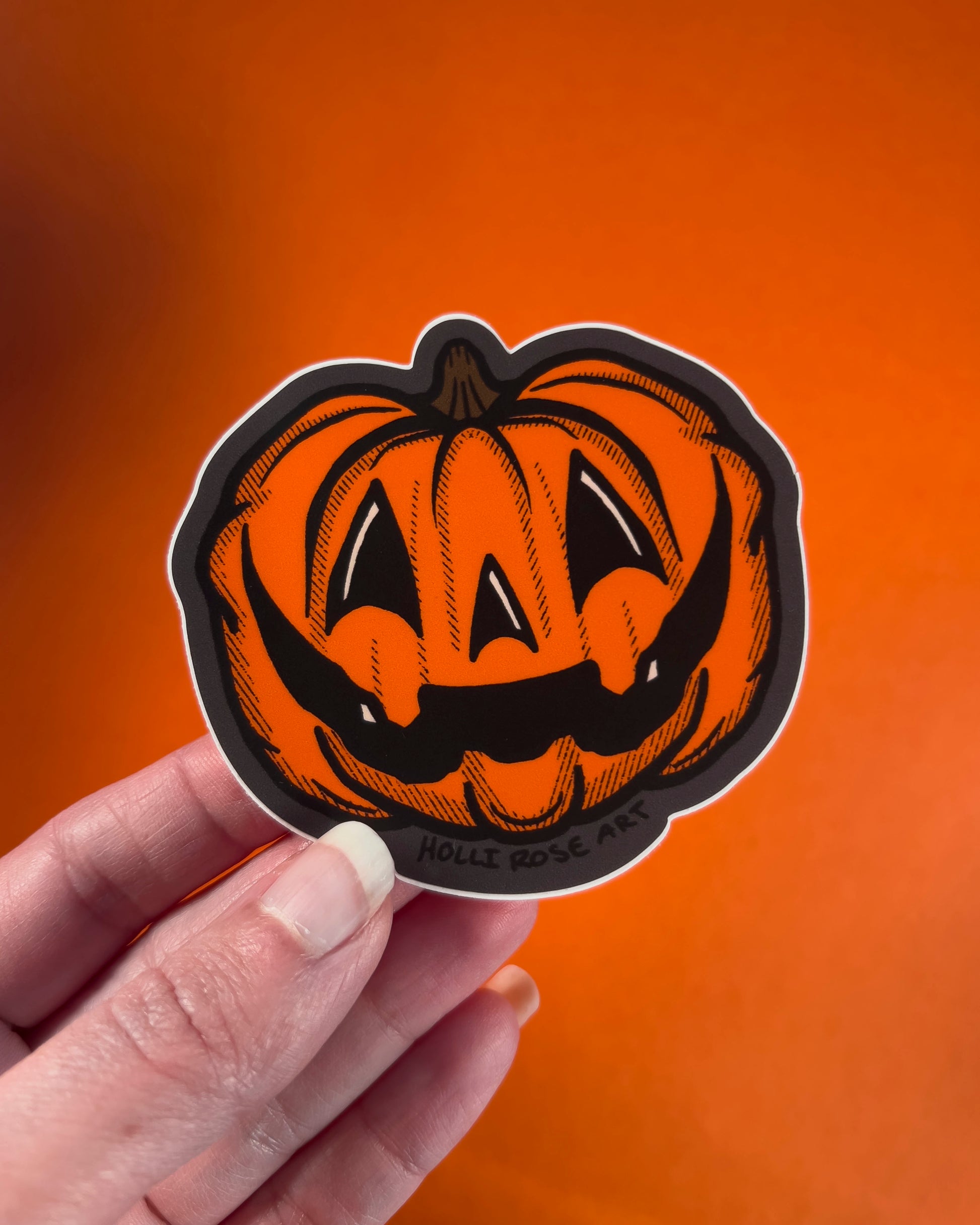 Hand holding a Halloween-themed pumpkin sticker against an orange background