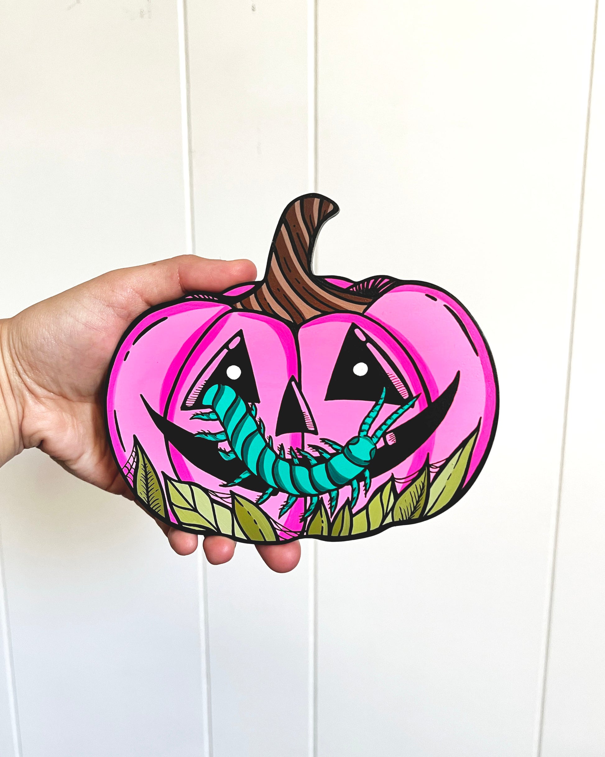 Hand holding a pink pumpkin with a cartoonish face and green worm design against a white background