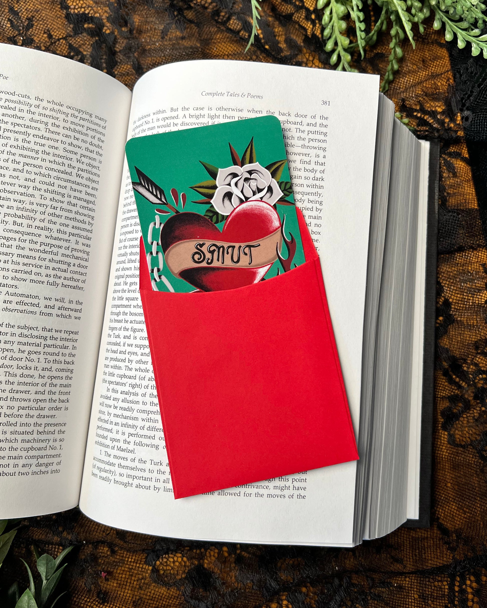 A colorful bookmark with a heart and floral design, featuring the word 'Smut', in a red bookmark sleeve  placed in an open book.