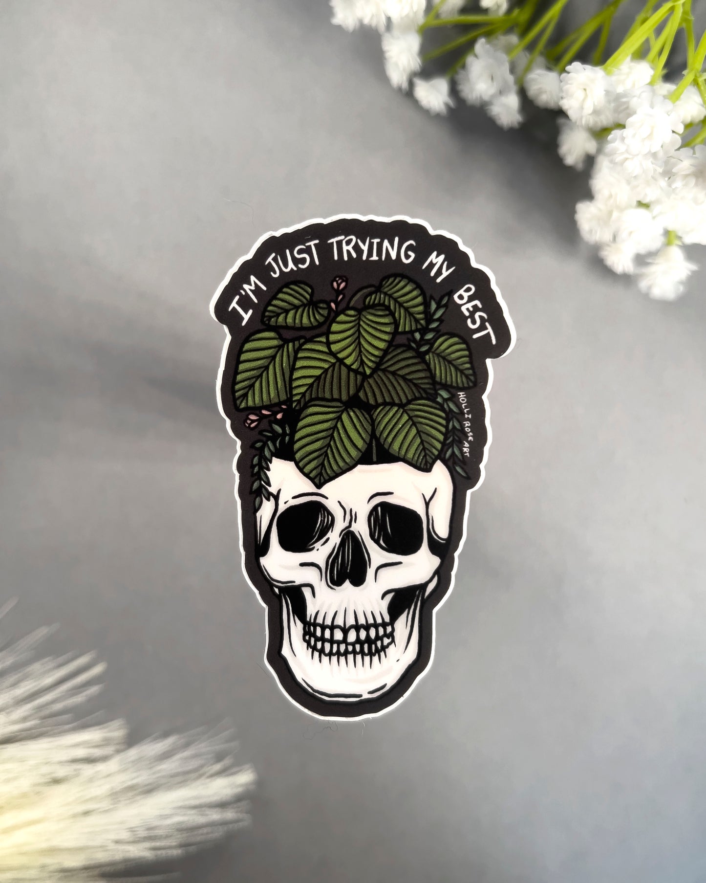Skull sticker with leaves and text 'I'm just trying my best' on a gray background with white flowers.
